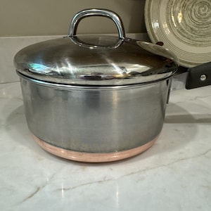 May include: A stainless steel saucepan with a matching lid and a copper-colored base. The pot features a single black handle attached with a metal rivet. The lid has a polished silver finish and a curved handle on top for easy lifting and handling.
