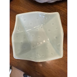 May include: A square, light mint green ceramic bowl with a glossy finish and a subtle, irregular, faceted shape. The interior surface features a pattern of small, raised, circular dots distributed across the base and sides of the vessel.