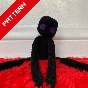 May include: Black crocheted plush toy with purple eyes, resembling a character from the video game Minecraft. The toy is sitting on a red fluffy surface.