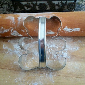 May include: A silver metal cookie cutter shaped like a bear. The cutter is on a wooden surface covered in white flour.