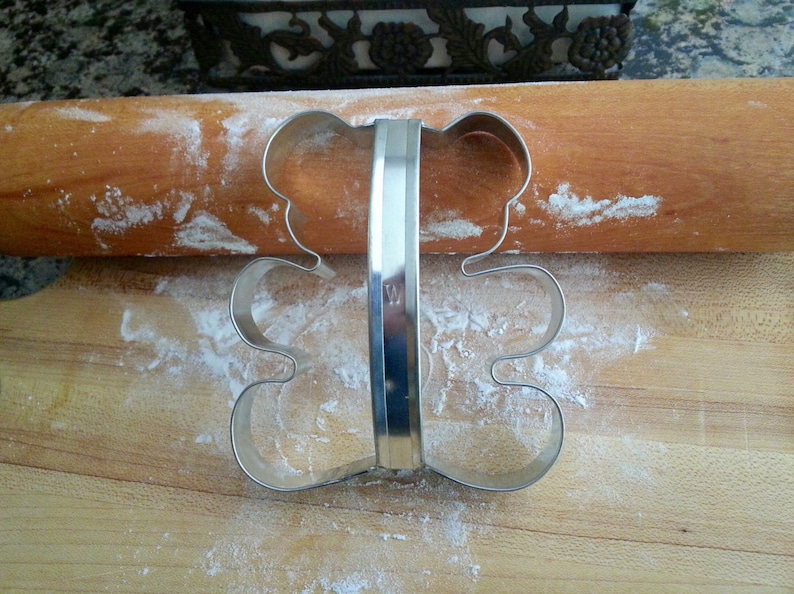 May include: A silver metal cookie cutter shaped like a bear. The cutter is on a wooden surface covered in white flour.