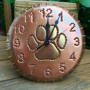 May include: Copper clock with a paw print design and black hands. The numbers 1 through 12 are around the edge of the clock.