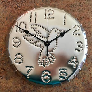 May include: A silver metal clock with a hummingbird design. The clock face has black hands and black numbers. The numbers are made of small metal circles.