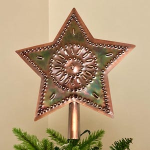 May include: A copper-colored star-shaped Christmas tree topper with a decorative design. The star has a central pattern and is mounted on a copper-colored pole. The star is approximately 10 inches wide.