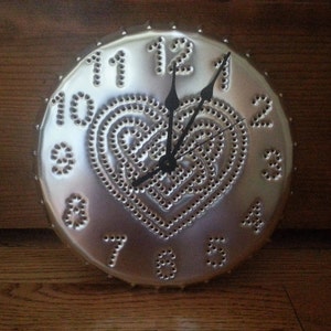 May include: A silver metal clock with a heart-shaped design and Roman numeral hour markers. The clock has a black hour and minute hand.