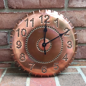 May include: A copper-colored wall clock with black hands and a circular design. The clock face features numbers created from small, evenly spaced holes. The clock is mounted on a red brick wall.
