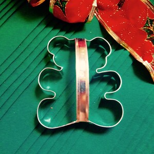 May include: A silver metal cookie cutter shaped like a bear. The cutter has a handle and is on a green surface.