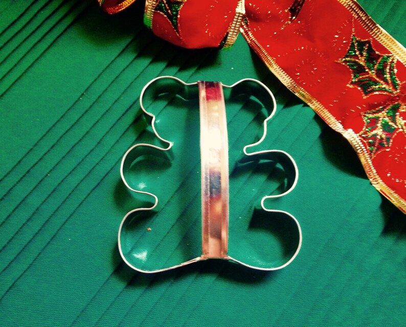 May include: A silver metal cookie cutter shaped like a bear. The cutter has a handle and is on a green surface.