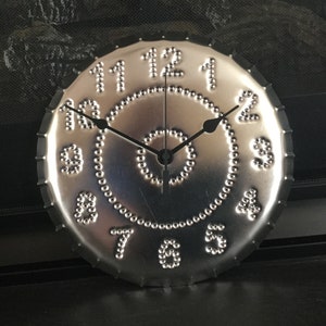 May include: A silver metal clock with black hands and a circle of raised dots around the edge. The numbers 1 through 12 are embossed on the face of the clock.