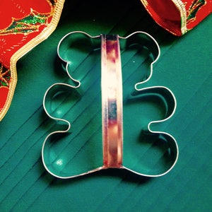 May include: A metal cookie cutter shaped like a teddy bear with a handle. The cookie cutter is on a green surface.