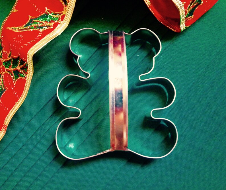 May include: A metal cookie cutter shaped like a teddy bear with a handle. The cookie cutter is on a green surface.