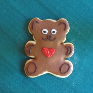 May include: A brown teddy bear cookie decorated with white eyes, a brown nose, and a red heart. The cookie is on a teal green surface.