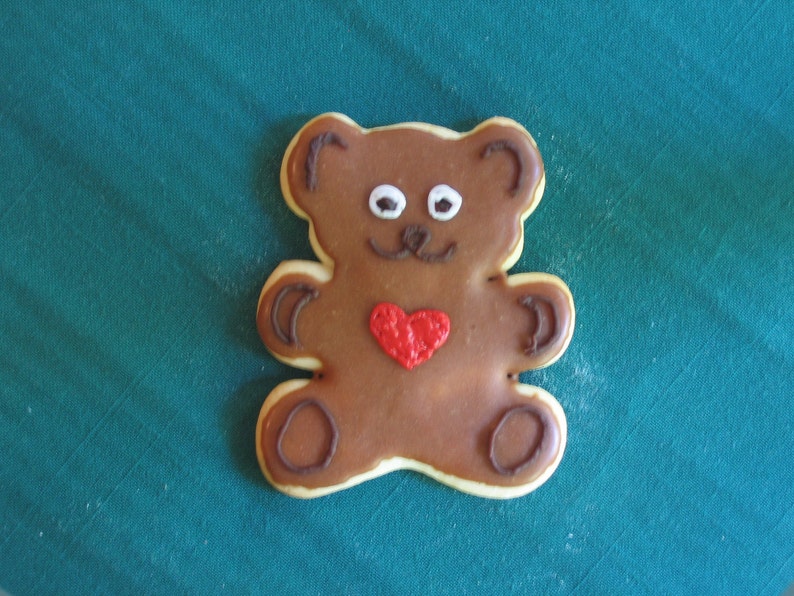 May include: A brown teddy bear cookie decorated with white eyes, a brown nose, and a red heart. The cookie is on a teal green surface.