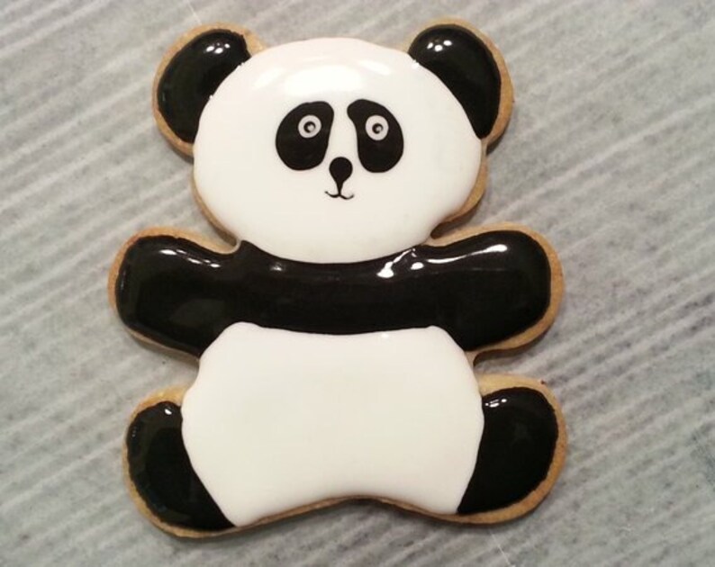 May include: A black and white panda bear cookie decorated with icing. The cookie is shaped like a bear with black ears, arms, and legs.