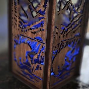 May include: Square wooden lantern featuring a mountain and bear silhouette design. The dark brown wood has blue accents. The design is cut out, allowing light to pass through. The lantern is a decorative item.