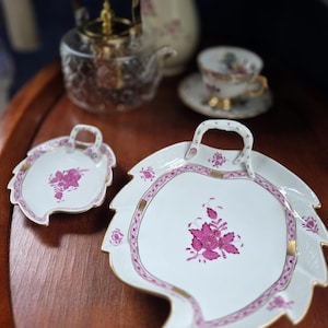 May include: White porcelain serving dishes with a leaf shape and a handle. The dishes have a pink floral design and gold trim. A glass teapot and teacup are in the background, suggesting a tea set.