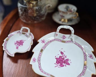 May include: White porcelain serving dishes with a leaf shape and a handle. The dishes have a pink floral design and gold trim. A glass teapot and teacup are in the background, suggesting a tea set.