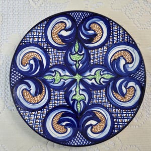 May include: A round, decorative ceramic plate with a detailed blue and white floral design. The plate features a central green cross-like pattern, surrounded by swirling blue and white motifs with brown accents. The rim is black.