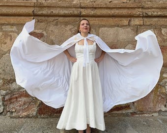 May include: A white dress and cape ensemble. The dress features a fitted bodice and a flowing skirt. The cape, draped over the shoulders, creates a dramatic silhouette. The person is standing against a stone wall, creating a striking contrast.