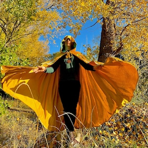 May include: A person wearing a yellow and green hooded cape stands in a forest with fall foliage. The cape is flowing in the wind and the person is looking up at the sky.