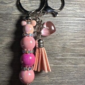 Puede incluir: Llavero de cuentas rosa y blanco con un dije de corazón rosa y un borla rosa. El llavero está unido a un anillo de llave plateado.