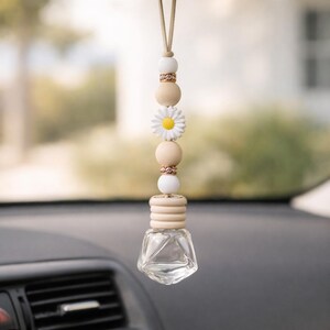 May include: Car air freshener with a clear, geometric-shaped glass bottle and a daisy charm. The air freshener is suspended by a beige cord and features wooden beads and gold accents. The car interior is visible in the background.