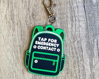 May include: A black and green backpack-shaped keychain with the text "TAP FOR EMERGENCY CONTACT". The keychain has a silver clasp and is on a wooden surface. The backpack design includes a zipper and flower details.