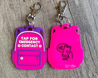 May include: Two pink backpack-shaped keychains with silver clasps. One keychain has the text "TAP FOR EMERGENCY CONTACT" in white. The other keychain has a graphic of a phone and hand with the letters "NFC".
