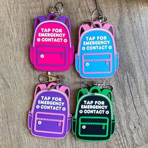May include: Four brightly coloured backpack keyrings. Each keyring displays the text "TAP FOR EMERGENCY CONTACT" in white. The backpacks are in pink, blue, purple, and black, with contrasting trim and a small flower detail.