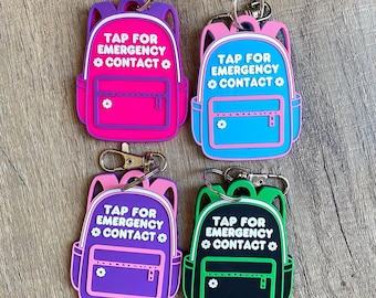 May include: Four colorful backpack-shaped keychains. Each keychain features the text "TAP FOR EMERGENCY CONTACT" in white. The backpacks are in pink, blue, purple, and black, with contrasting trim and a small flower detail.