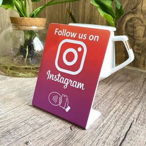 May include: White sign with a red, orange, and purple gradient. The sign says "Follow us on Instagram" with the Instagram logo and a stylized image of a phone with a wave symbol.
