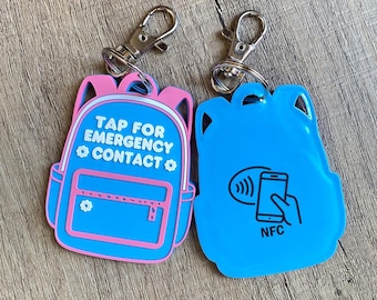 May include: Two backpack-shaped keychains. One is pink and blue with the text "TAP FOR EMERGENCY CONTACT". The other is blue with a phone and NFC symbol. Both have silver clasps.