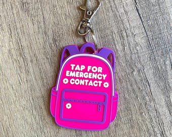 May include: A pink backpack-shaped keychain with purple accents and a silver clasp. The keychain has the text "TAP FOR EMERGENCY CONTACT" in white. A small white flower is on the front of the backpack.