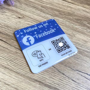 May include: Blue and white square sticker with the text "Follow us on Facebook" and a Facebook logo. The sticker also has a QR code with the text "SCAN QR CODE ID: 9CC3AA" and a symbol of a phone with the text "NFC TAP PHONE"
