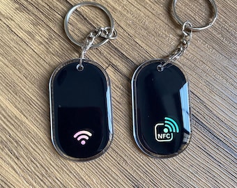 May include: Two oval-shaped black keychains with silver metal rings and chains. The left keychain features a colorful Wi-Fi symbol icon. The right keychain displays a white and teal NFC icon with the text NFC inside a rounded square outline.