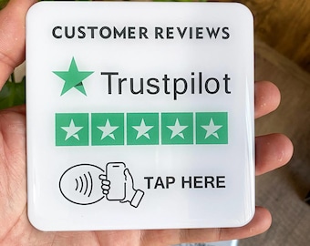 May include: White sign with the text "CUSTOMER REVIEWS" and "Trustpilot" with five green stars. The sign also has a black and white illustration of a hand holding a phone with a wave symbol next to it and the text "TAP HERE".