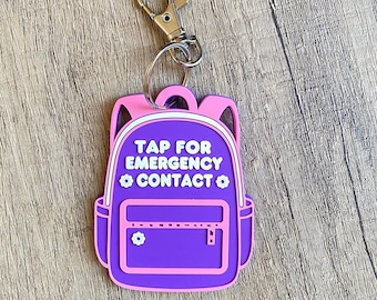 May include: A purple and pink backpack-shaped keychain with the text "TAP FOR EMERGENCY CONTACT". The keychain has a gold-colored clasp and a small flower detail on the front pocket.