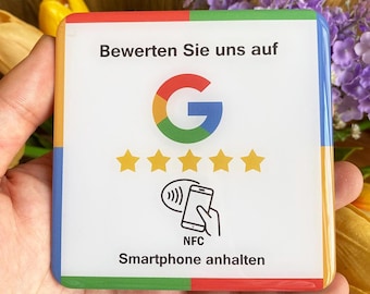 May include: Square white sign with colorful edges, featuring the Google logo, five gold stars, and a smartphone icon with the text "NFC Smartphone anhalten". The sign is held in a hand, with flowers in the background.