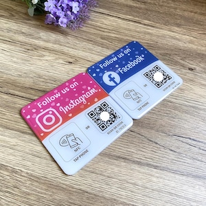 May include: Two square stickers with pink and blue backgrounds. The stickers have a white border and feature social media icons for Instagram and Facebook. Each sticker has a QR code and text that says "Follow us on" and "SCAN QR CODE".