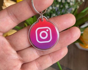 May include: A clear acrylic keychain with a silver metal ring. The keychain features a round charm with the Instagram logo in white on a gradient background of red, orange, and purple.