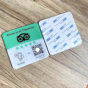 Könnte beinhalten: Ein rechteckiges, grün-weißes Schild mit dem Text "Review us on Tripadvisor" und einem QR-Code. Das Schild zeigt auch das TripAdvisor-Eulenlogo, Sterne und Anweisungen für NFC-Tap. Neben dem Schild befindet sich ein quadratisches Klebepad.