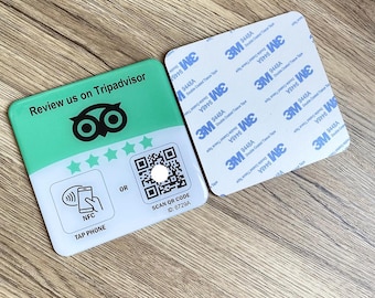 May include: A rectangular, green and white sign with the text "Review us on Tripadvisor" and a QR code. The sign also features the TripAdvisor owl logo, stars, and instructions for NFC tap. A square adhesive pad is next to the sign.
