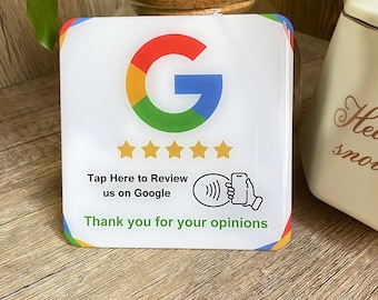 May include: White sign with the Google logo, five gold stars, and the text "Tap Here to Review us on Google". Below the text is a black and white icon of a phone with a wave symbol, indicating contactless payment. The sign also says "Thank you for your opinions".
