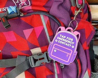 May include: A close-up of a colorful backpack with a geometric pattern in red, purple, and pink. A purple backpack-shaped keychain with the text "TAP FOR EMERGENCY CONTACT" is attached to the backpack.
