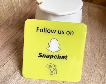 May include: A square, bright yellow sign with rounded corners. The sign reads "Follow us on Snapchat" in black text, with the Snapchat ghost logo. A smartphone icon with NFC symbol is at the bottom. A white ceramic container is in the background.