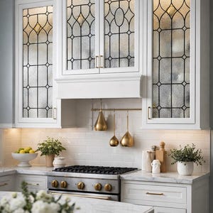May include: A bright, modern kitchen with white cabinetry and stained glass cabinet doors. The kitchen features a stainless steel gas range, gold-toned hardware, and decorative hanging kitchen utensils. A marble countertop and white subway tile backsplash complete the look.