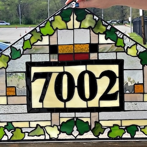 May include: A stained glass window with a triangular shape and a leafy border. The center of the window features a rectangular panel with the number 7002 in black lettering.