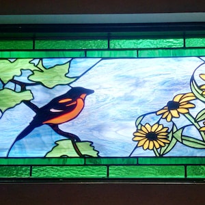 May include: A stained glass window depicting a bird perched on a branch with green leaves and yellow flowers. The bird is orange, black, and white. The background is blue and white.