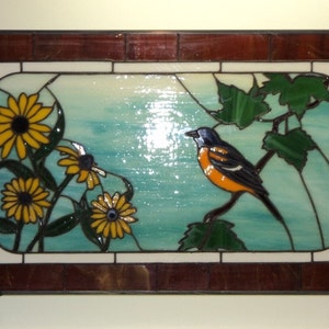 May include: A stained glass window depicting a bright orange and black bird perched on a green leafy branch. The bird is surrounded by yellow sunflowers and green leaves against a blue and white background.