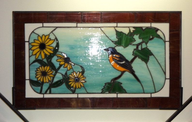 May include: A stained glass window depicting a bright orange and black bird perched on a green leafy branch. The bird is surrounded by yellow sunflowers and green leaves against a blue and white background.